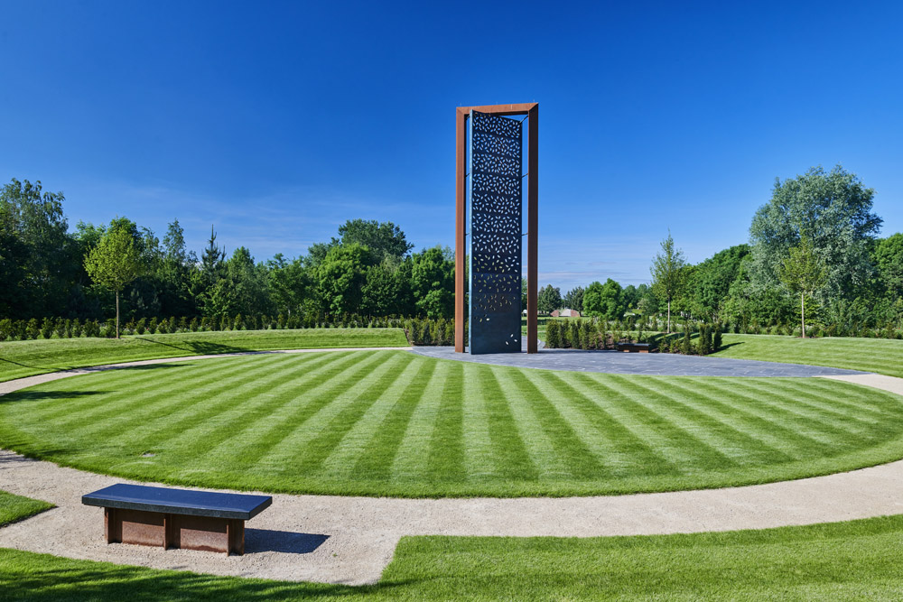Tall bronze and stone police memorial with perforated brass screen, leaf‑shaped apertures, surrounding trees, and reflective open lawn.